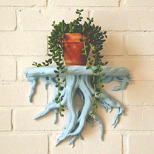 Może przedstawiać: Jasnoniebieska półka w kształcie korzeni drzewa, zamontowana na białej ceglanej ścianie. Na półce stoi terakotowa doniczka z zieloną rośliną zwisającą. Design półki przypomina korzenie drzewa, dodając naturalnego akcentu.