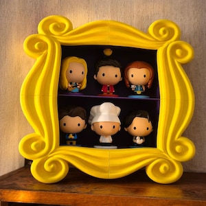 Puede incluir: Un marco de fotos amarillo con seis figuras de dibujos animados de personajes de la serie de televisión Friends. El marco es de color amarillo brillante y tiene un diseño decorativo en espiral. Las figuras están en dos estantes.