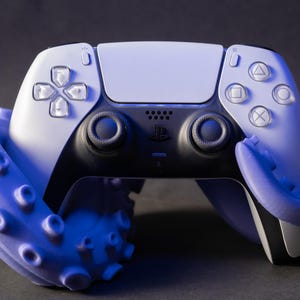 Peut inclure: Une manette PlayStation 5 blanche et noire avec un accessoire en forme de poulpe bleu. La manette a un design symétrique avec des boutons et des joysticks. L'accessoire poulpe entoure la manette.