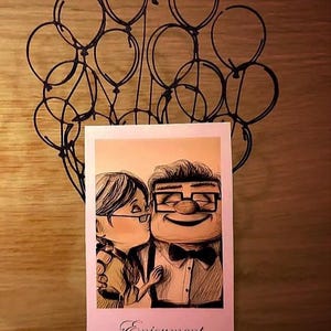 Pode incluir: Ilustração emoldurada de um casal se beijando, com a palavra "Enjoyment" na parte inferior. A ilustração é sustentada por uma escultura de arame preto de balões. O fundo é uma superfície de madeira.