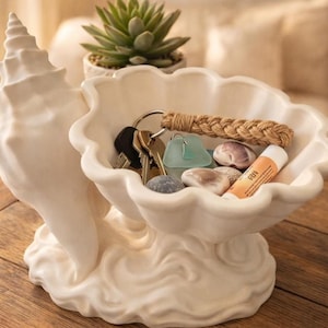 Könnte beinhalten: Eine cremefarbene, muschelförmige Dekoschale. Die Schale enthält Schlüssel, Meerglas und einen Lippenbalsam. Der Boden der Schale hat ein wellenartiges Design. Im Hintergrund befindet sich eine Sukkulentenpflanze.