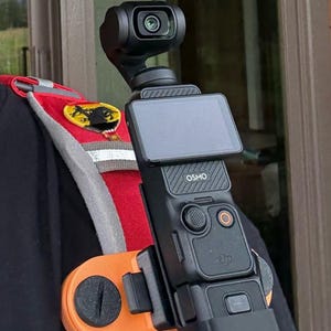 May include: A black Osmo Pocket camera with a built-in screen and buttons. The camera is mounted on an orange and black chest harness. The camera has a small lens and is designed for video recording.