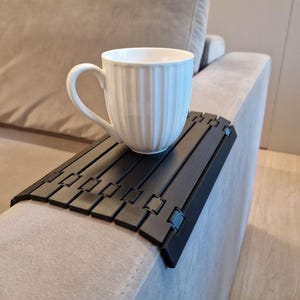 May include: A white ceramic mug with vertical ridges rests on a black, segmented sofa arm tray. The tray is designed to fit over the arm of a sofa, providing a stable surface for drinks or snacks.