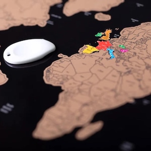 May include: A black world map with a scratch-off surface, revealing a tan color underneath. A white, teardrop-shaped object is on the left. Several European countries are highlighted in various colours, including red, yellow, and blue.