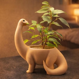 Può includere: Un vaso a forma di dinosauro beige con un lungo collo e coda, contenente una pianta verde. Il vaso ha un'ampia apertura per la pianta ed è realizzato in un materiale liscio. La pianta ha foglie verdi e steli marroni.