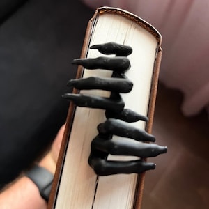 Pode incluir: Um marcador de livros preto em forma de mão, agarrando as páginas de um livro. O marcador de livros é feito de um material escuro e está posicionado na lateral do livro, com os dedos estendidos para baixo.