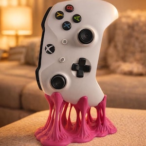 Pode incluir: Um controle Xbox branco é exibido em um suporte rosa, semelhante a chiclete. O controle possui botões coloridos e um direcional preto. O suporte tem uma aparência texturizada e derretida, criando uma exibição única.
