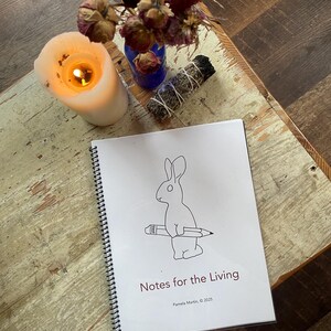 May include: A lit white candle, dried flowers in a blue vase, and a bundle of sage sit on a wooden table. A spiral-bound notebook with a rabbit holding a pencil and the text "Notes for the Living" is in the foreground.