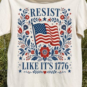 May include: A cream-colored short-sleeve t-shirt featuring a graphic print with the text RESIST at the top and LIKE IT'S 1776 at the bottom. The design includes a central American flag surrounded by red and blue floral patterns and small stars.
