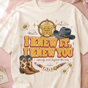 以下が含まれることがあります： クリーム色のTシャツで、'THIS AIN'T MY FIRST RODEO'と書かれた金色の保安官バッジ、青いカウボーイハット、茶色の柄入りカウボーイブーツ、デイジーの花、'TS5'と書かれたビーズの紐のグラフィックプリントが施されています。テキストは'I KNEW IT, I KNEW YOU'および'infinity and beyond the eras'と書かれています。