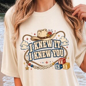 以下が含まれることがあります： カウボーイハット、投げ縄、そして「I KNEW IT, I KNEW YOU」という文字がプリントされたクリーム色のTシャツ。デザインには小さな雲、星、SWIFTIEと綴られたビーズの紐、T、S、5と書かれた2つの積み木が含まれています。