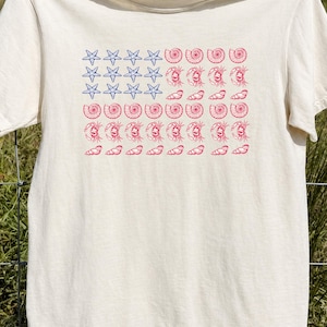 May include: A cream-colored short-sleeve t-shirt featuring a graphic print arranged in a flag pattern. The design consists of blue starfish in the upper left corner, with rows of red seashells and crabs forming the remaining stripes of the flag.