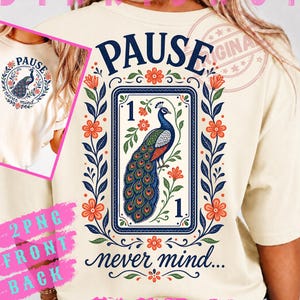 May include: A cream-colored t-shirt featuring a graphic design of a peacock inside a rectangular frame with the number 1 at the top and bottom. The text PAUSE is arched above the frame, and never mind is written below in a cursive script.