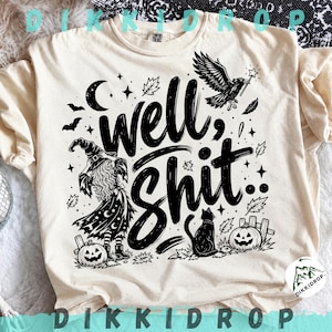 Könnte beinhalten: Ein cremefarbenes Kurzarm-T-Shirt mit einem schwarzen Grafikdruck und dem Text "well, SHIT". Das Design zeigt eine Hexe, eine fliegende Krähe mit Zauberstab, eine schwarze Katze, zwei Kürbisse, Fledermäuse, Sterne, eine Mondsichel und Blätter.