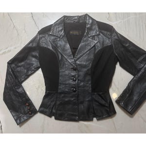May include: A black jacket featuring a combination of leather and fabric panels. The garment has a collared neckline, a three-button front closure, and long sleeves with three buttons at each cuff. The design includes a fitted waistline.