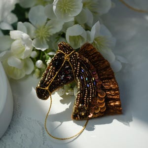 May include: A handcrafted horse head brooch, featuring a detailed design of brown and gold beads and sequins. The brooch has a gold-coloured bridle and is set against a backdrop of white flowers.