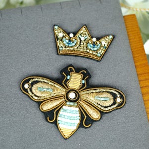 May include: Two embroidered patches: a gold and teal crown and a bee. The crown is gold with teal and white accents. The bee has gold wings, a teal and white striped body, and black detailing. Both are on a grey surface.