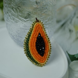 May include: A beaded papaya brooch. The brooch features a detailed design of a papaya slice, with orange, green, and black beads. The edges are outlined with gold-colored beads, creating a decorative accessory.