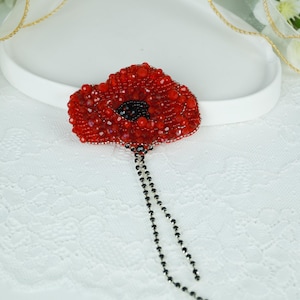 Può includere: Spilla artigianale a forma di fiore di papavero rosso con perline, con centro nero e fili pendenti di perline nere. La spilla è realizzata con piccole perline lucenti, su sfondo bianco.