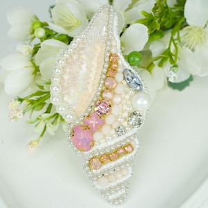 May include: A seashell-shaped brooch, embellished with pearls, sequins, and various pink and clear gemstones. The brooch has a white base and gold accents. The background features white flowers.