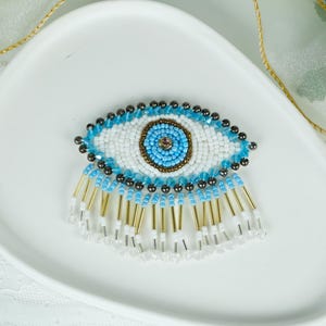 Puede incluir: Broche con forma de ojo con cuentas con fondo blanco, iris azul y flecos dorados y transparentes. El ojo tiene un diseño de cuentas azules y blancas con un pequeño centro rosa. El broche está sobre un plato ovalado blanco.