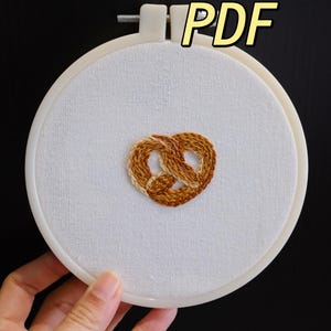 Peut inclure: Un cercle à broder avec un tissu blanc brodé d'un motif de bretzel en tons bruns. Le mot "PDF" est dans un encadré jaune en haut. Le cercle est tenu par une main sur fond noir.