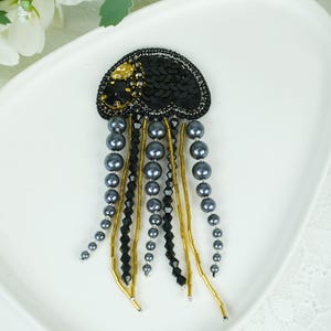 May include: A handcrafted brooch in the shape of a jellyfish. The black sequined body is adorned with gold and black beads. Gray pearl strands and gold beaded strands form the tentacles, creating a unique accessory.