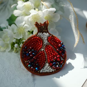 Puede incluir: Broche artesanal de granada, con un diseño de cuentas rojas vibrantes con detalles blancos e iridiscentes. El broche está intrincadamente detallado, mostrando una forma de fruta realista. Un accesorio único.