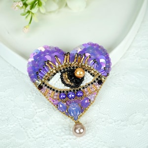 May include: A heart-shaped brooch with an eye design. The brooch is adorned with purple sequins, gold and black beads, and a dangling pearl. The eye has a white iris, black pupil, and a gold bead. The brooch is on a white surface.