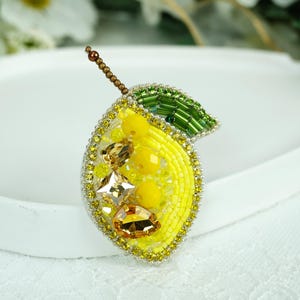 May include: A handmade lemon brooch, crafted with yellow beads and embellished with clear and amber crystals. It features a green beaded leaf and a brown stem. This decorative brooch is suitable for adding to clothing or accessories.