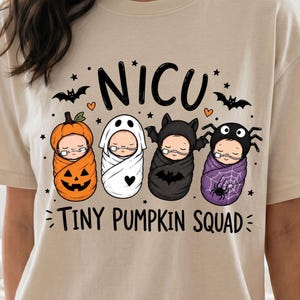 Può includere: Una t-shirt beige a maniche corte con una grafica di quattro neonati in fasce vestiti da zucca, fantasma, pipistrello e ragno. Il testo NICU è scritto sopra e TINY PUMPKIN SQUAD sotto, decorato con piccole stelle, cuori e pipistrelli.