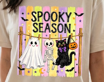 Puede incluir: Una camiseta beige con un gráfico de un tendedero con un fantasma, un esqueleto y un gato negro sosteniendo una calabaza. El texto SPOOKY SEASON está impreso encima, sobre un fondo de rayas verticales en tonos pastel y estrellas.
