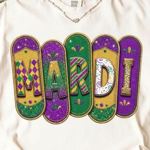 May include: Cream-coloured t-shirt featuring the word "MARDI" in a vibrant, glittery design. The letters are in gold, green, and purple, with decorative borders. The shirt is made of a soft material.
