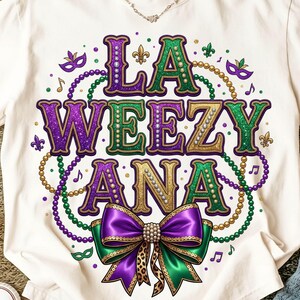 Può includere: T-shirt bianco con un design grafico colorato. Il design presenta le parole "LA WEEZY ANA" in viola, verde e oro, circondate da perline, note musicali e un fiocco con un centro a stampa leopardata. La maglietta ha uno scollo tondo.