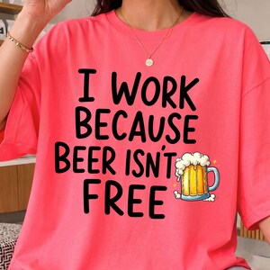 以下が含まれることがあります： 鮮やかなコーラルピンクの半袖Tシャツ。黒い太字で「I WORK BECAUSE BEER ISN'T FREE」とプリントされています。「FREE」という文字の横には、黄金色の液体と白い泡が入ったビールジョッキのグラフィックイラストが描かれています。
