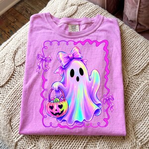 Puede incluir: Una camiseta rosa claro con el gr&aacute;fico de un fantasma hologr&aacute;fico que lleva un lazo grande y sostiene un cubo de calabaza lleno de dulces. El dise&ntilde;o est&aacute; encerrado en un borde festoneado rosa brillante con lazos peque&ntilde;os a los lados.