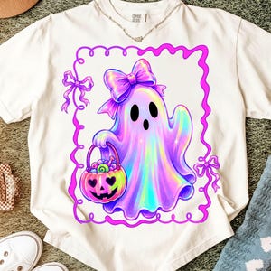 Puede incluir: Una camiseta blanca con el gr&aacute;fico de un fantasma hologr&aacute;fico con un gran lazo morado, sosteniendo un cubo de dulces con forma de calabaza. El dise&ntilde;o est&aacute; encerrado en un borde rosa ondulado con peque&ntilde;os lazos en las esquinas, en el frente.