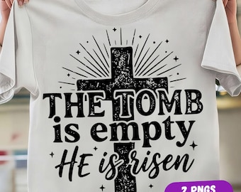 May include: A white short-sleeved t-shirt featuring a black graphic of a cross with a distressed texture. The text reads THE TOMB is empty HE is risen, surrounded by radiating lines and small star accents. The design is centered on the chest.