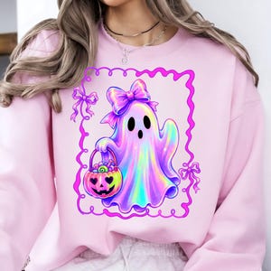 Puede incluir: Una sudadera rosa claro con un gr&aacute;fico central de un fantasma de estilo hologr&aacute;fico con un lazo grande en la cabeza, sosteniendo un cubo de dulces de calabaza. El dise&ntilde;o est&aacute; encerrado en un marco rectangular rosa brillante con bordes ondulados.