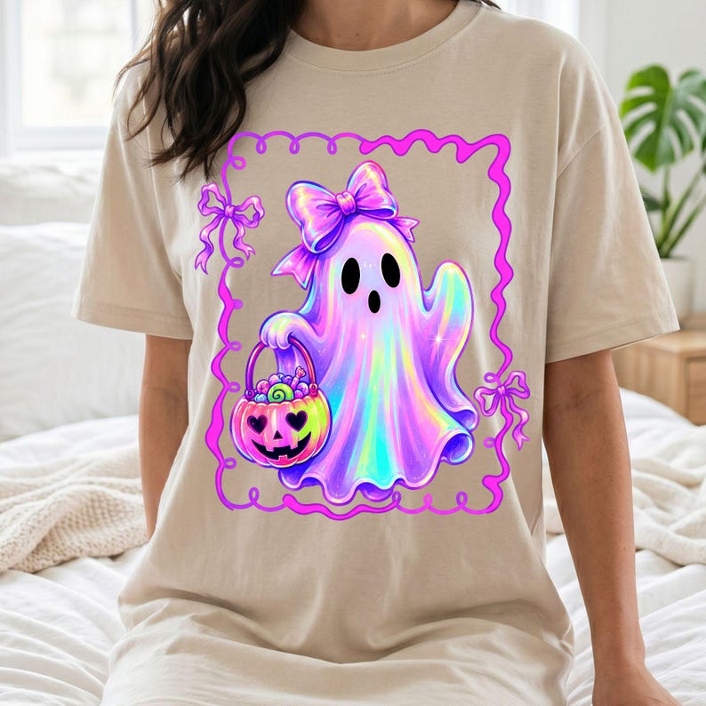 Puede incluir: Una camiseta beige con el gr&aacute;fico de un fantasma hologr&aacute;fico con un lazo grande en la cabeza, sosteniendo un cubo de calabaza lleno de dulces. El dise&ntilde;o est&aacute; encerrado en un borde rectangular ondulado de color rosa brillante.