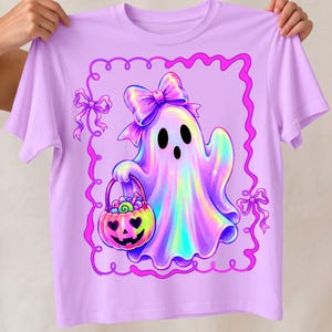 Puede incluir: Una camiseta de color morado claro con el gr&aacute;fico de un fantasma hologr&aacute;fico que lleva un lazo grande en la cabeza. El fantasma sostiene un cubo de dulces con forma de calabaza lleno de golosinas. El dise&ntilde;o est&aacute; dentro de un marco rosa.