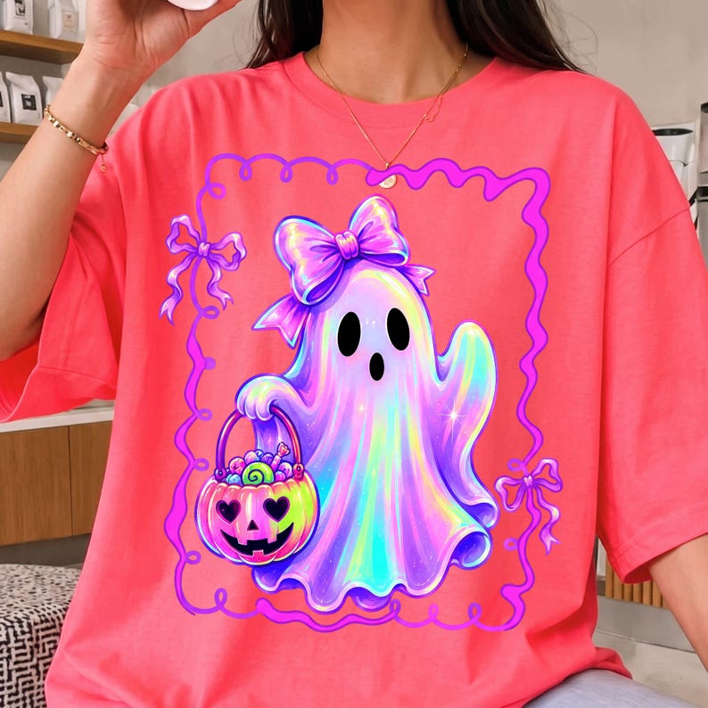 Puede incluir: Una camiseta de color rosa ne&oacute;n brillante con un gr&aacute;fico de un fantasma hologr&aacute;fico con un lazo grande en la cabeza. El fantasma sostiene un cubo de dulces en forma de calabaza lleno de golosinas, todo dentro de un marco decorativo morado.