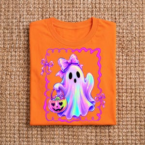 Puede incluir: Una camiseta de color naranja brillante con un gr&aacute;fico de un fantasma de estilo hologr&aacute;fico con un lazo morado en la cabeza. El fantasma sostiene un cubo de dulces con forma de calabaza, todo dentro de un borde rectangular rosa ondulado.
