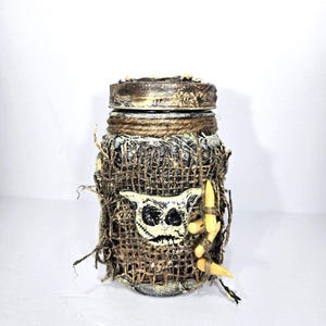 Pode incluir: Um frasco decorativo com uma aparência rústica e artesanal. O frasco é envolto em serapilheira e adornado com um emblema em forma de caveira e detalhes em forma de osso. A tampa é pintada com um acabamento metálico e o frasco é enrolado com barbante.