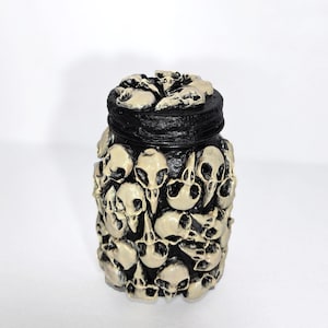 May include: A black jar with a lid, adorned with numerous off-white skulls. The skulls are closely packed, forming a textured pattern. The jar is probably ceramic or a similar material, with detailed skull features.