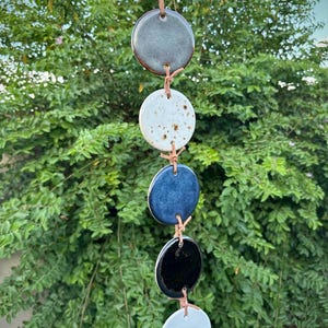 May include: A vertical wall hanging featuring five circular ceramic discs connected by thin tan leather cords. The discs are finished in varying glazes: a metallic grey, a speckled white, a deep blue, a glossy black, and a solid light grey.
