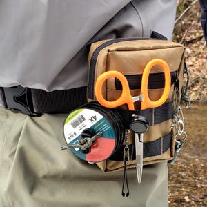 Puede incluir: Una bolsa de aparejos de pesca de color canela sujeta a un cinturón, con tijeras naranjas, una bobina de hilo de pescar y otras herramientas. La bolsa tiene múltiples compartimentos y correas. La bobina de hilo de pescar está etiquetada "6.4 LB".