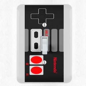 May include: A white and black light switch cover designed to resemble a classic Nintendo controller. The cover features a directional pad, red buttons, and the Nintendo logo. The switch itself is white.