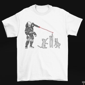 May include: White t-shirt featuring a humorous graphic of a Predator character aiming a laser at three cats. The Predator is in gray armor, and the cats are in various poses. The laser is red.