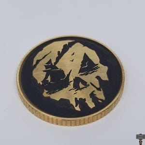 May include: A round, brass-colored coin with a black skull and crossbones design. The coin features a detailed engraving of a ship sailing near an island. The coin's edge has a ridged texture.